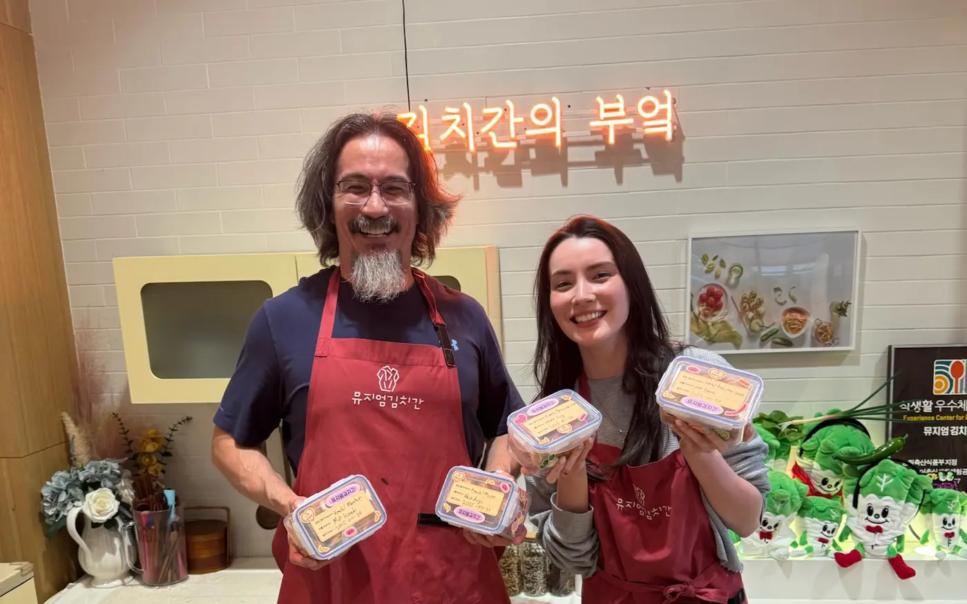 John and Fiona smiling in red aprons while holding containers of kimchi