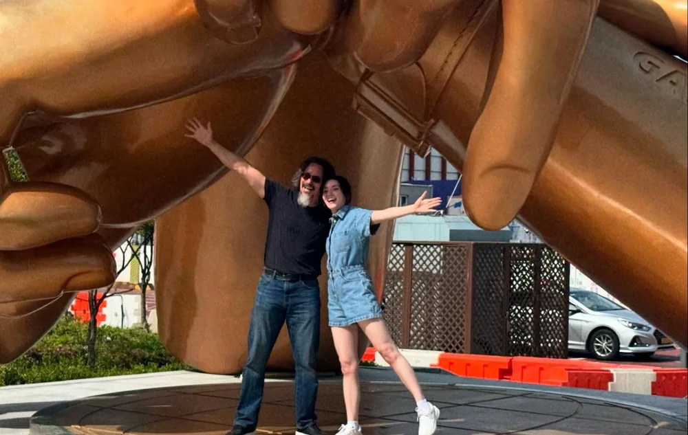 John and Fiona posing beneath a large bronze hand sculpture during their 2025 trip to Korea