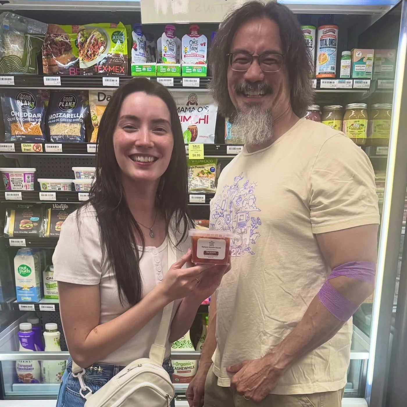Fiona Daub and John Daub holding a jar of their mat kimchi in front of the cooler at Village Foods & Pharmacy in Bryan, Texas